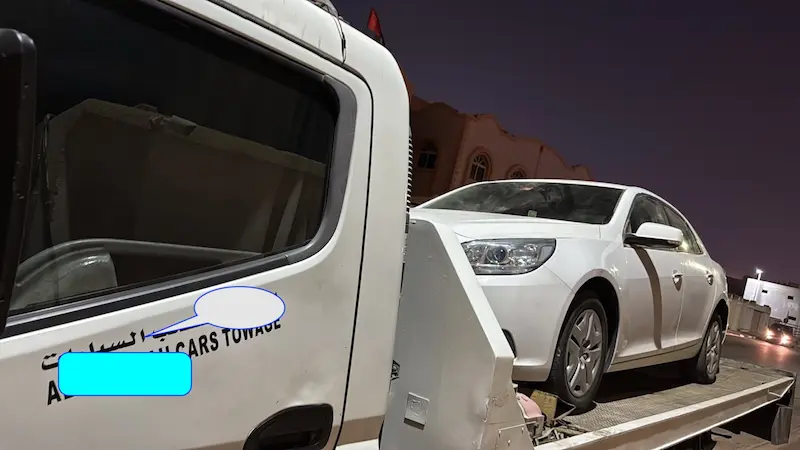 Side view of a white sedan on a flatbed recovery truck before the move from Dasman to Industrial Area 5